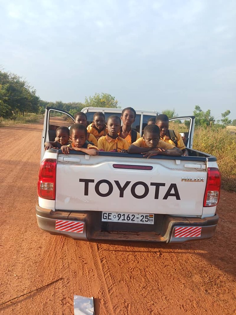 Eine Gruppe von Kindern, alle in gelben Uniformen, sitzen auf dem Rücksitz eines weißen Toyota-Pickups auf einer unbefestigten Straße.