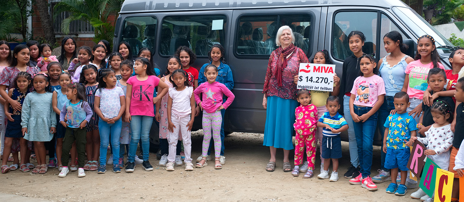 Eine große Gruppe von Kindern und einer älteren Frau posiert vor einem dunklen Transporter. Die Frau hält ein Schild mit der Aufschrift "MIVA" und einem Betrag, was auf eine Spendenaktion hindeutet. Die Szene strahlt eine Atmosphäre der Dankbarkeit und Gemeinschaft aus, wahrscheinlich im Rahmen eines Hilfsprojekts.